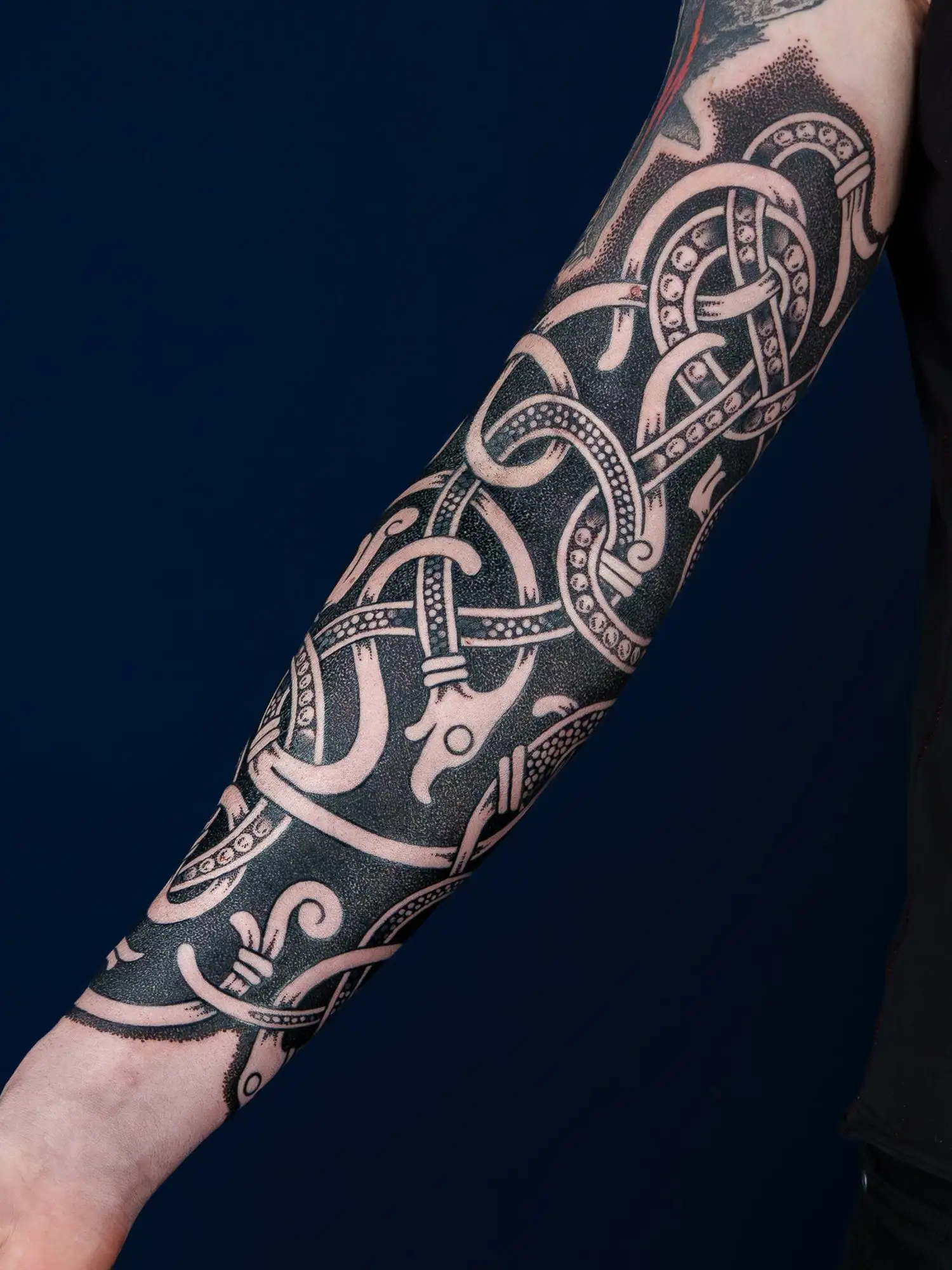 Ein Arm mit einem schwarzen, detaillierten Tattoo im nordischen Stil, das auf einem dunkelblauen Hintergrund sichtbar ist.