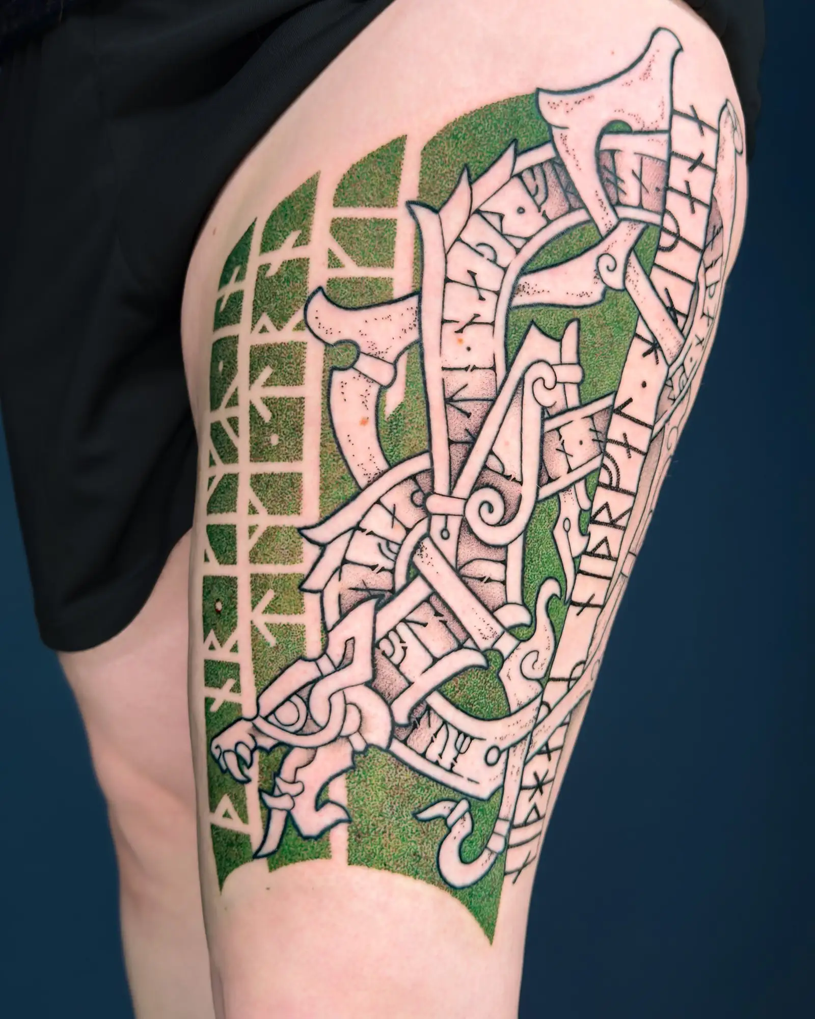 Ein detailliertes grünes und weißes Wikinger-Tattoo auf einem Oberschenkel, das einen Drachenkopf und Runen zeigt.
