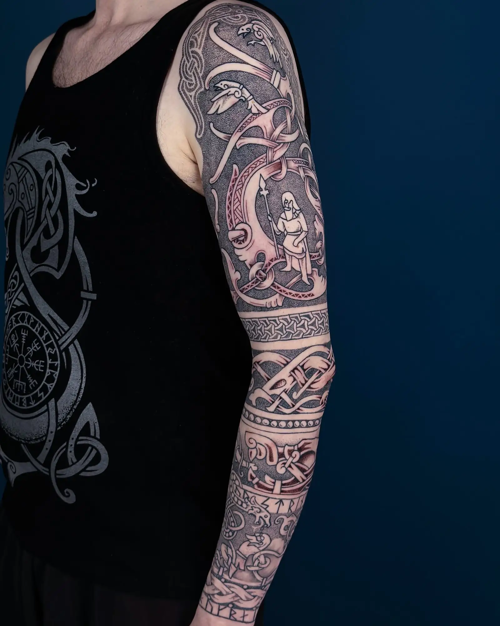 Arm mit einem aufwendigen Wikinger-Sleevetattoo, das Runen, Tiere und eine Figur mit Speer zeigt.