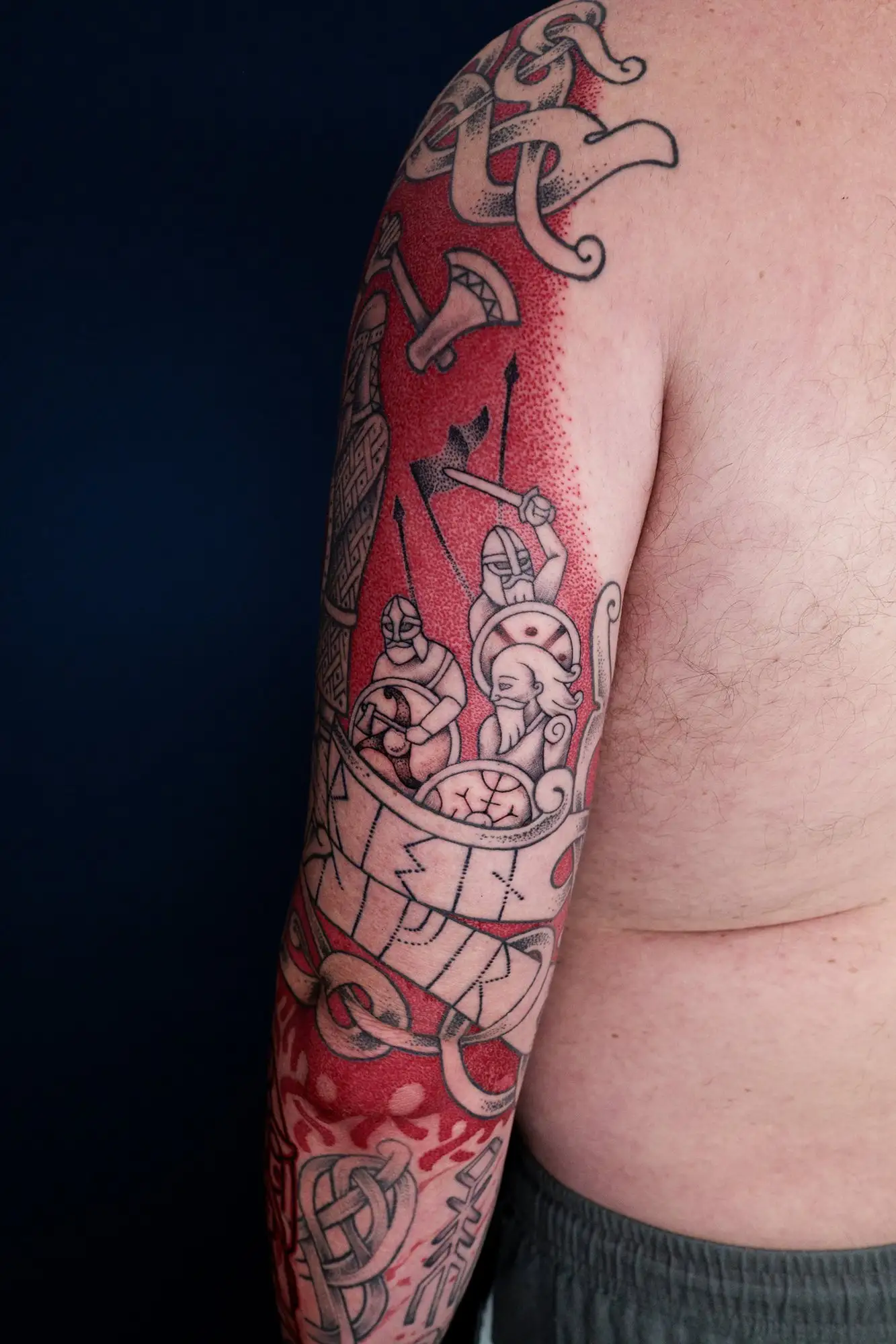 Viking-Tattoo auf einem Arm mit Kriegern in einem Langschiff, rotem Hintergrund und nordischen Motiven.